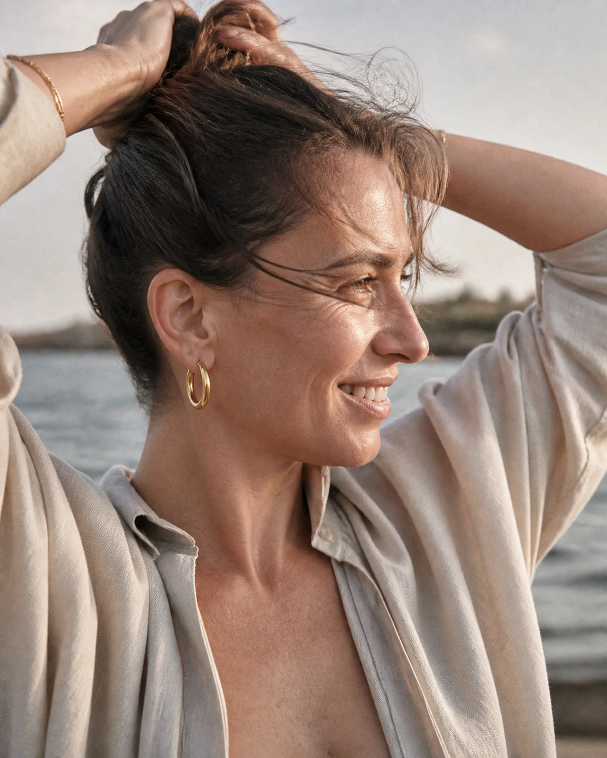 Woman by the water wearing a gold MAREA Everyday Hoop, tying her hair up
